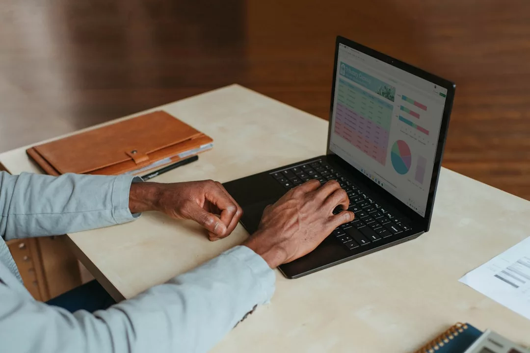 Person working at a desk using a laptop displaying charts and graphs, with a brown folder and pen beside them—a glimpse into the modern lifestyle where productivity meets fashion and sophistication.