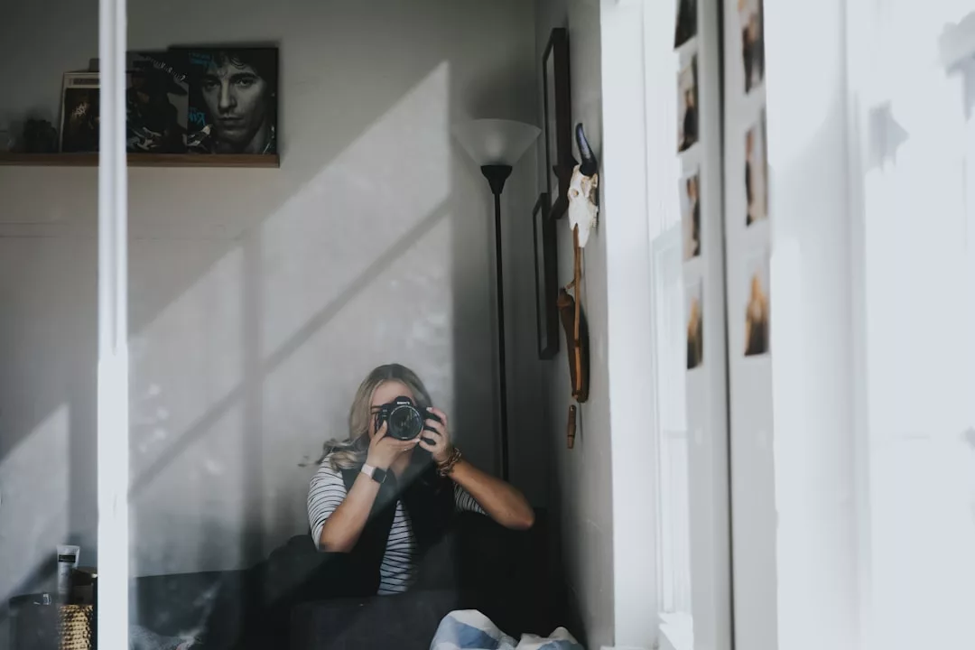 A person with blonde hair sits indoors, capturing their reflection in a mirror amid stylish wall decorations and photos, giving the room a touch of luxury lifestyle.