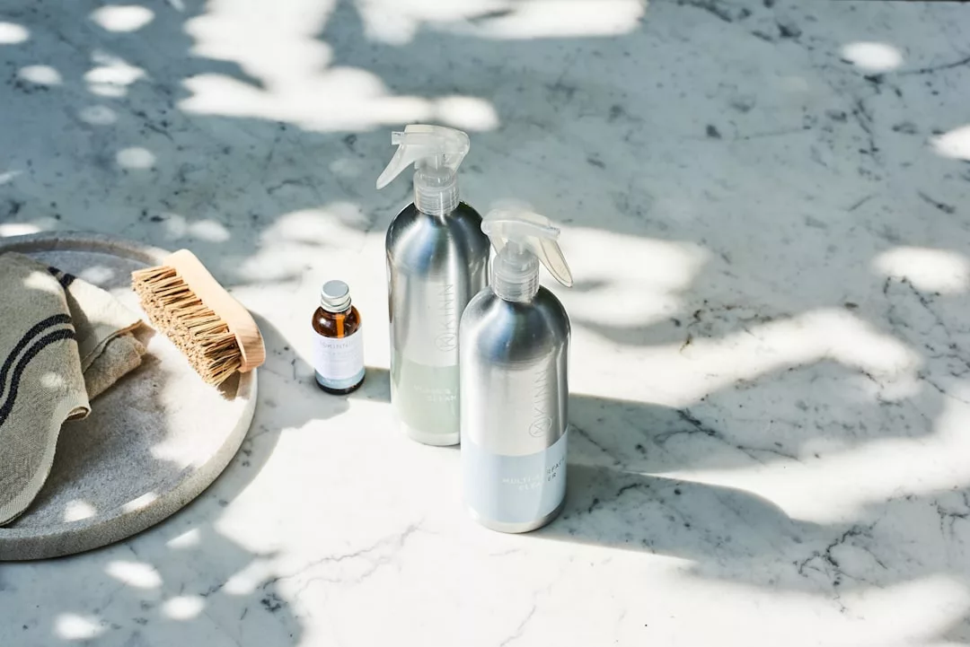 Two aluminum spray bottles, a small brown glass bottle, a wooden scrub brush, and a folded towel are arranged on a sunlit marble surface with dappled shadows, creating an effortless sense of luxury.
