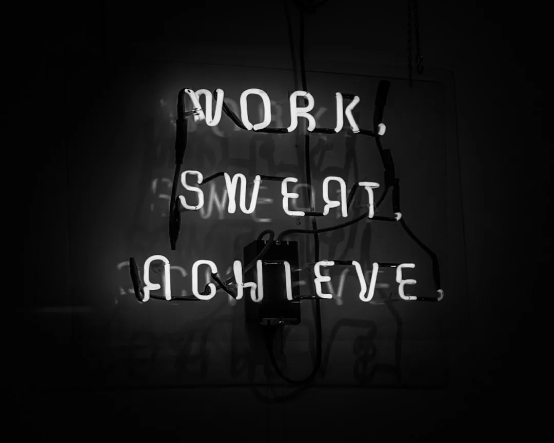 A neon sign glows with the words "WORK, SWEAT, ACHIEVE" against a dark background, inspiring a bold lifestyle of determination and ambition.