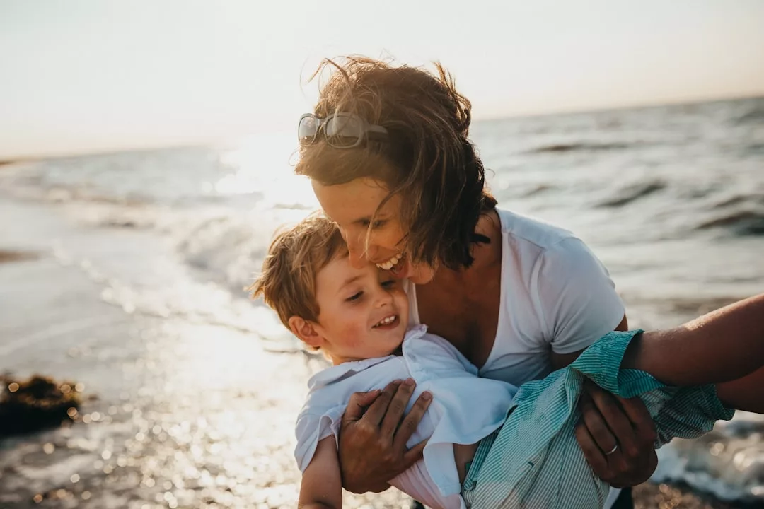 An adult holding a smiling child near the shoreline at sunset, both in light-colored clothing, enjoying a luxury lifestyle with ocean waves in the background.
