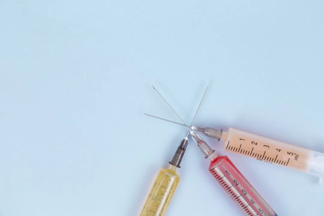 Three syringes with yellow, red, and pink liquids meet at the center on a light blue background, evoking a bold Glamesia aesthetic perfect for those who appreciate unique fashion and lifestyle visuals.