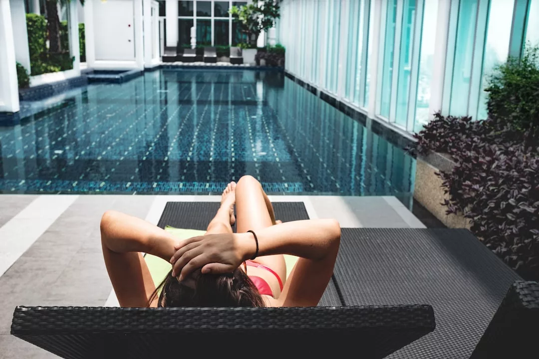 A person in swimwear reclines on a lounge chair beside an indoor swimming pool, embracing a luxury lifestyle with hands resting behind their head.