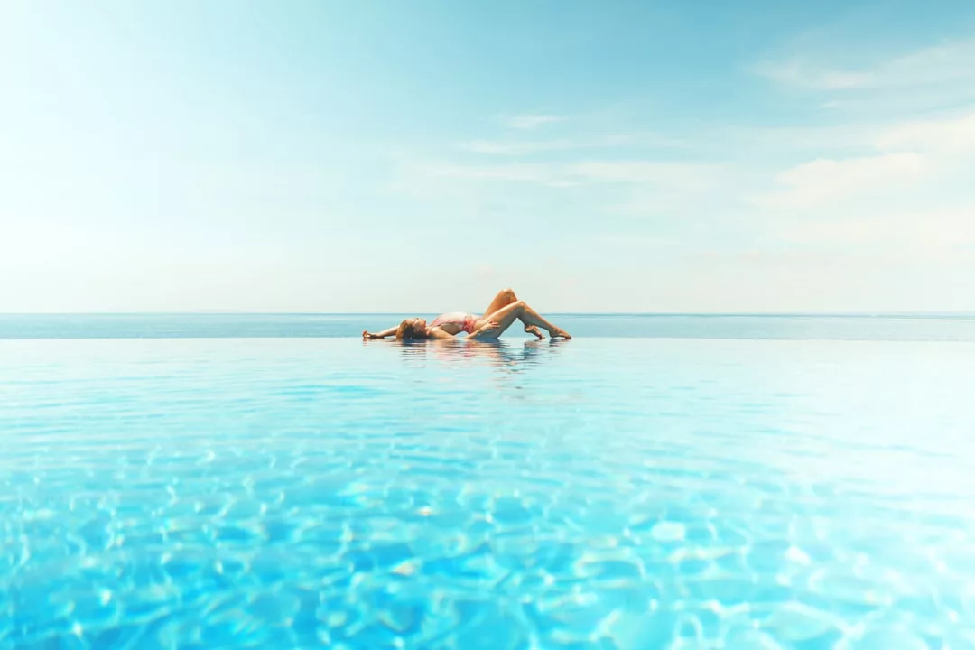 A person lies on their back at the edge of a luxury infinity pool overlooking a calm sea under a clear blue sky.