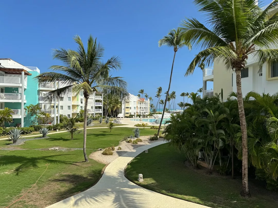 A paved walkway winds through a landscaped garden with palm trees, leading to white resort buildings and a pool under a clear blue sky—perfect for enjoying a luxe lifestyle.