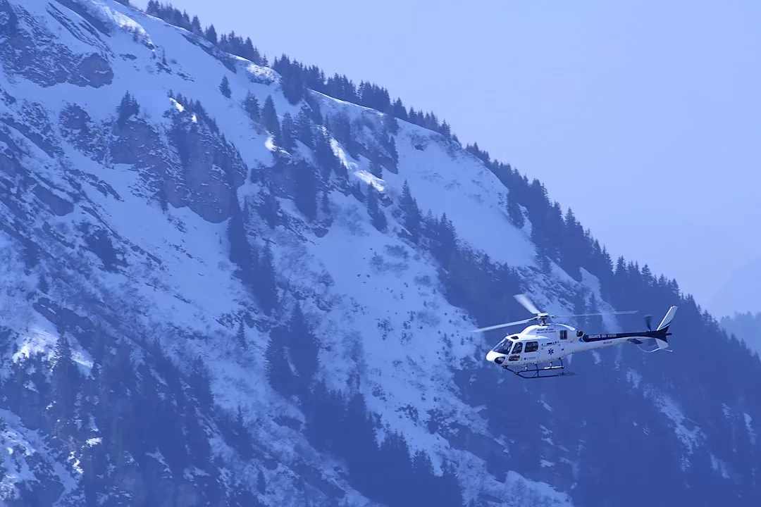 A white helicopter flies in front of a snow-covered mountain slope dotted with evergreen trees, capturing a scene of adventure and luxe lifestyle.