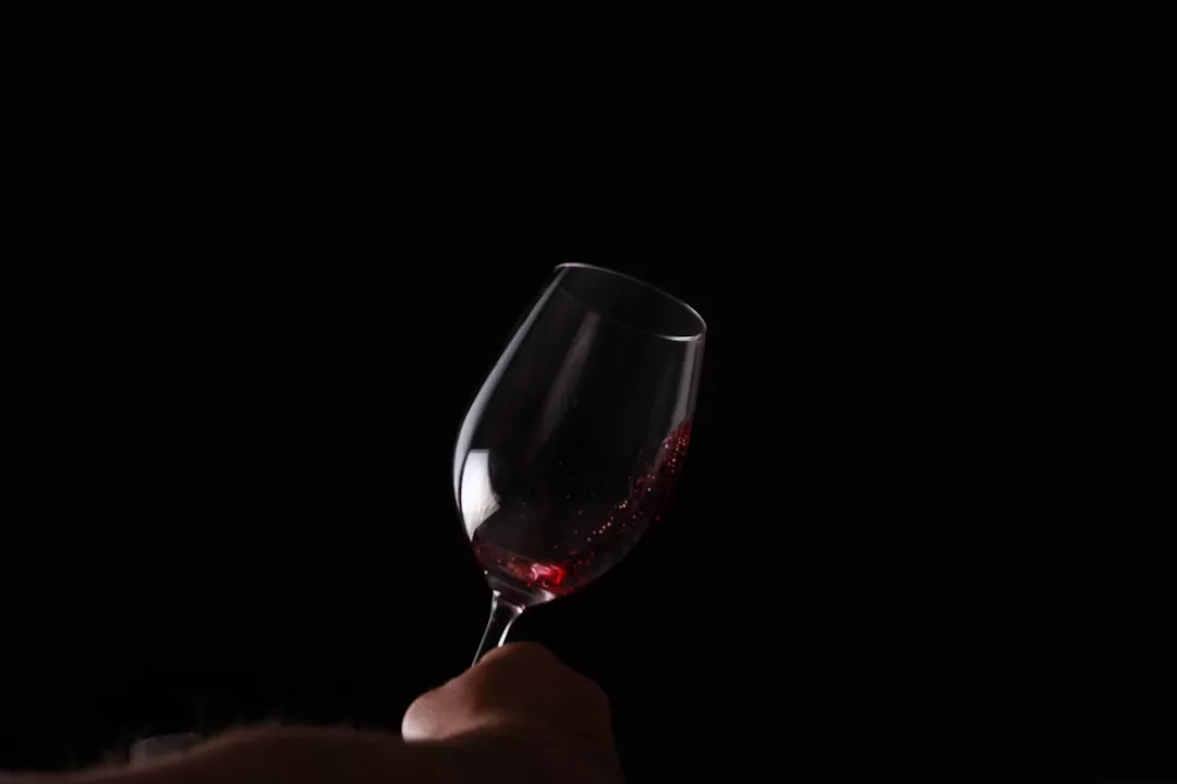 A hand holds a tilted wine glass with a small amount of red wine, exuding luxury against a plain black background.