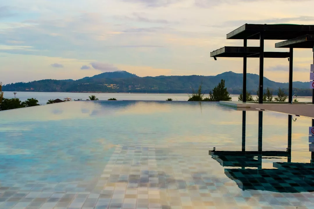 Infinity pool reflecting a modern Glamesia structure, with mountains and water visible in the distance under a cloudy sky at sunset, exuding fashion-forward luxury.