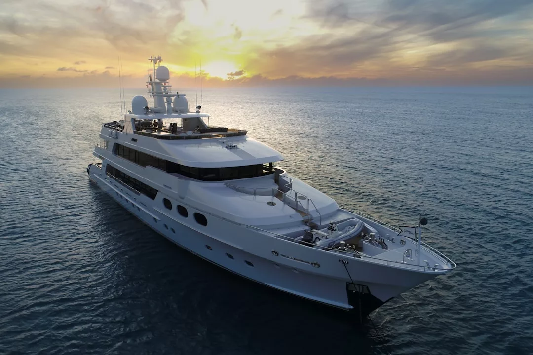 A large white Glamesia luxury yacht is anchored on calm ocean water at sunset, with the sun setting behind partial clouds on the horizon, offering a taste of the ultimate Laxary lifestyle.