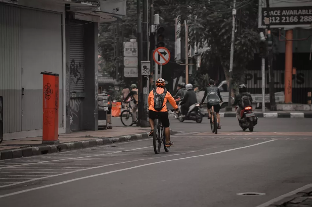 A cyclist in a luxury orange jacket rides on a city street, with other cyclists and a motorcyclist visible ahead.