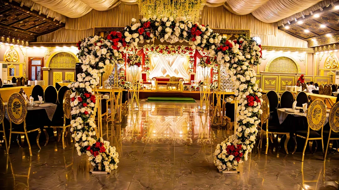 A lavishly decorated banquet hall by Glamesia features a floral archway of white and red flowers, gold chairs, ornate walls, and a stage with a red and gold sofa for true luxury celebrations.