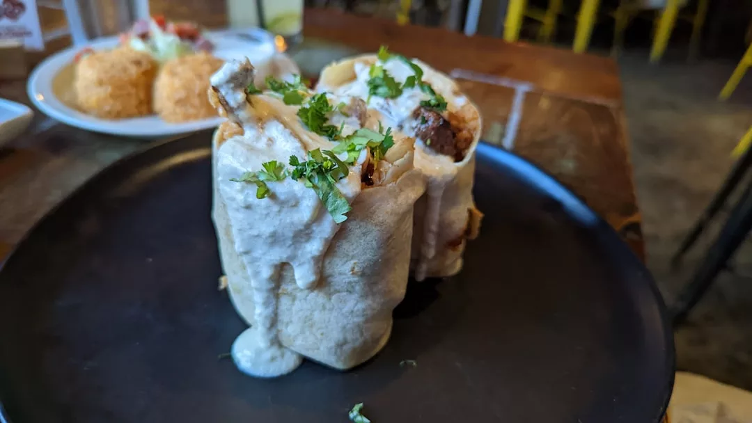 A close-up of a stuffed tortilla wrap topped with creamy white sauce and chopped herbs, served on a dark plate in a luxury restaurant setting.