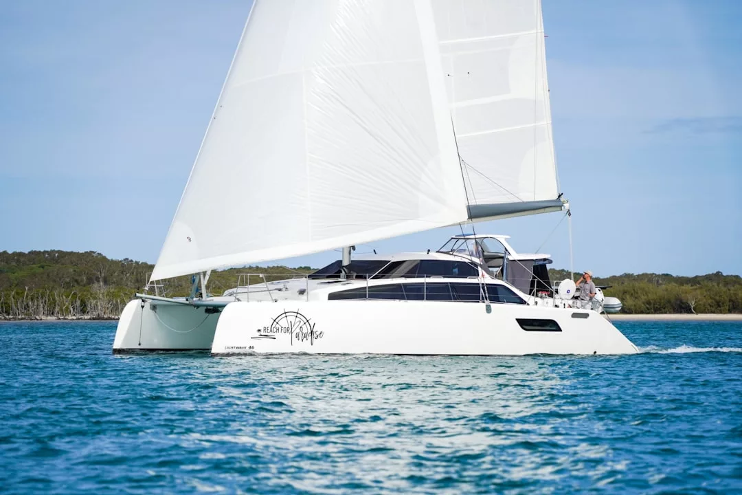 A luxury white catamaran sailboat with raised sails glides on blue water near a forested shoreline under a clear sky, offering the ultimate lifestyle experience.