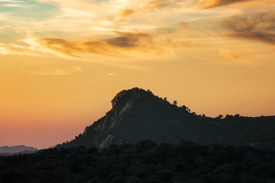 A rugged hilltop covered with trees is silhouetted against an orange and yellow sky at sunset, with wispy clouds above—a scene radiating natural luxury.