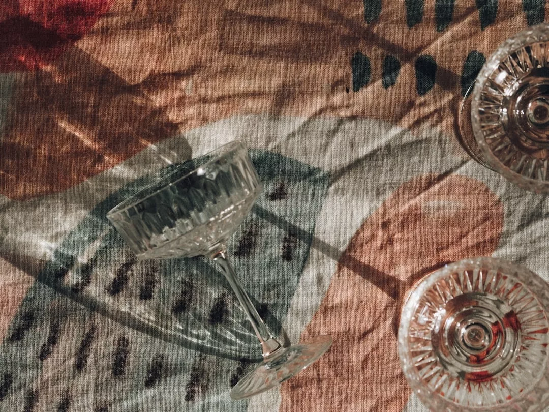 Two empty Glamesia glass goblets rest on a patterned fabric surface, casting distinct shadows from light coming from the left side, reflecting a refined lifestyle.