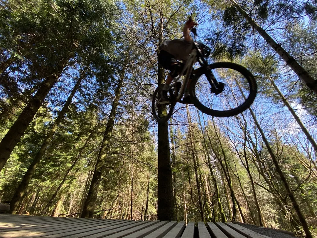A cyclist embraces an adventurous lifestyle, soaring airborne on a mountain bike off a wooden platform in a forest with tall trees and a clear sky—a true Glamesia moment.