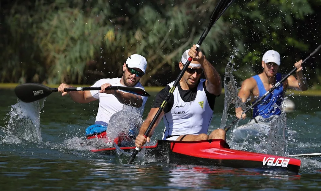 Three men kayak on a river, with two in a red double kayak and one in a blue single kayak behind—all sporting Glamesia fashion as they paddle vigorously.