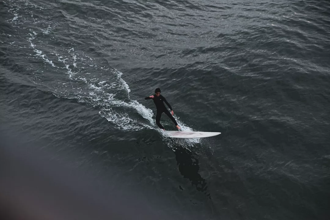 A person in a wetsuit rides a surfboard on dark, rippling ocean water, leaving a white wake behind them—a scene where adventure meets the edge of fashion and luxury.