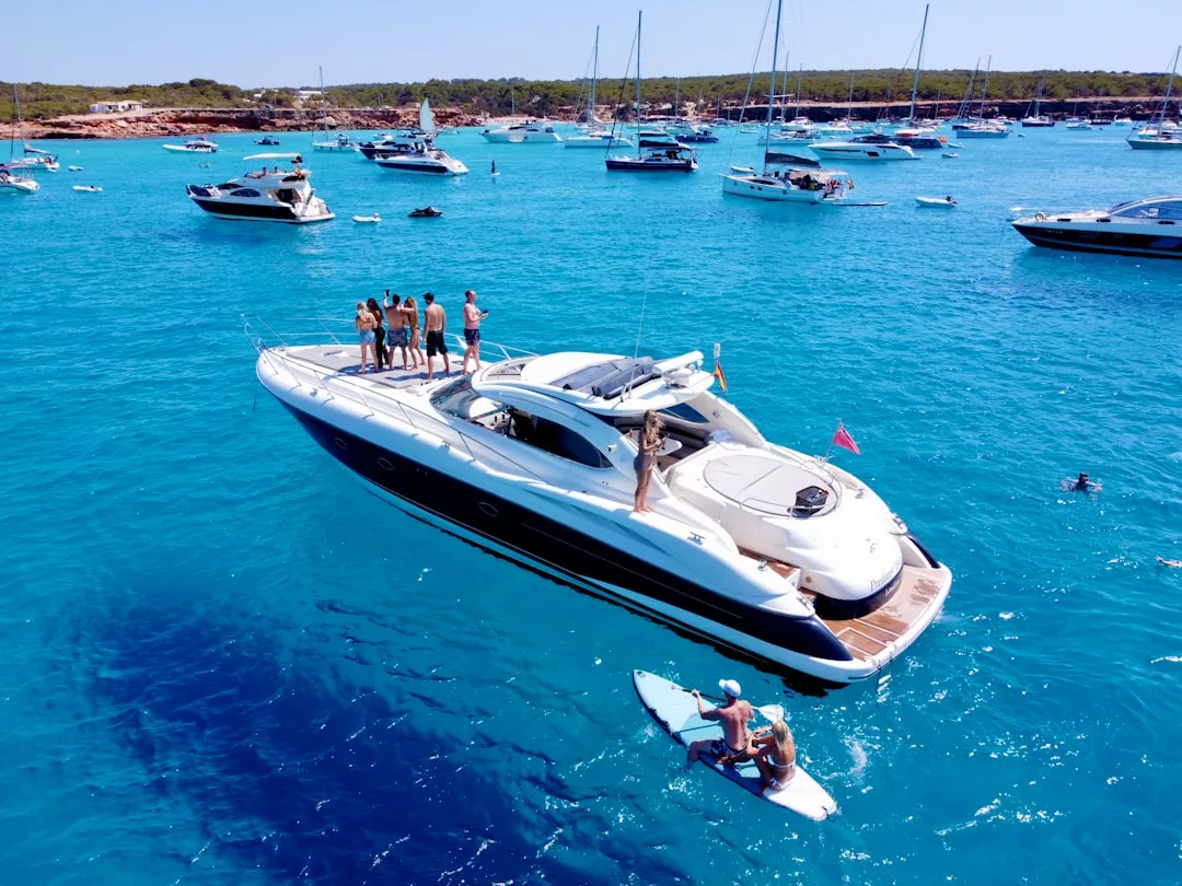 A white yacht with several people on board and one person paddling on a board in clear blue water, surrounded by other boats and a distant shoreline—a perfect scene of luxury lifestyle and relaxation.