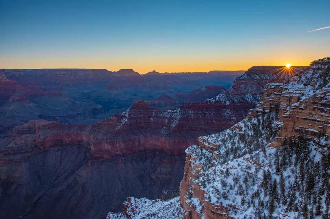 The sun sets over the snow-dusted cliffs and layered rock formations of the Grand Canyon, casting a luxury glow under a clear sky.