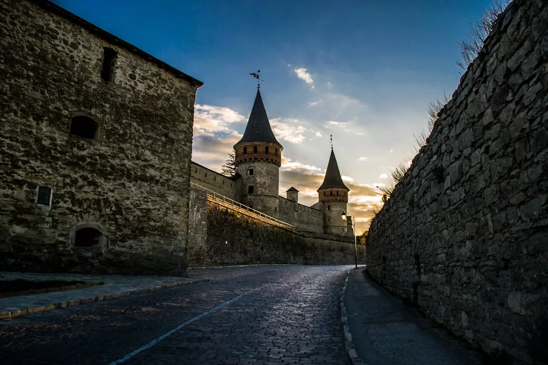 A cobblestone road leads to a luxury historic stone fortress with conical towers at sunset, flanked by high stone walls.