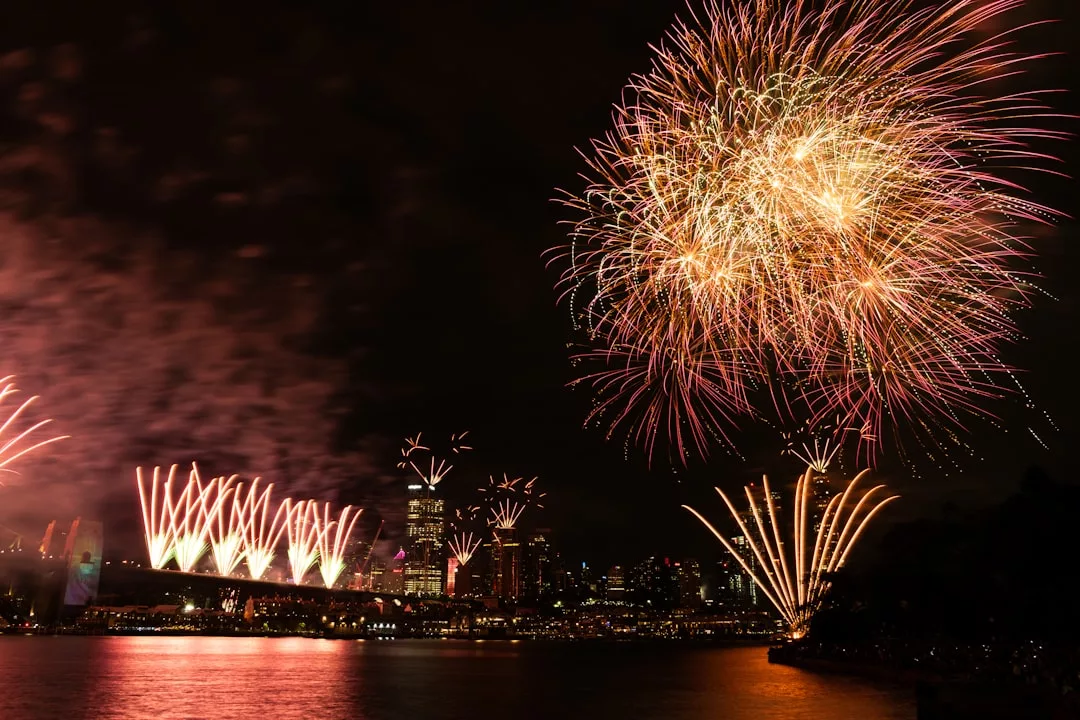 Fireworks light up the night sky over a city skyline, reflecting on the water below and creating a scene of pure luxury.