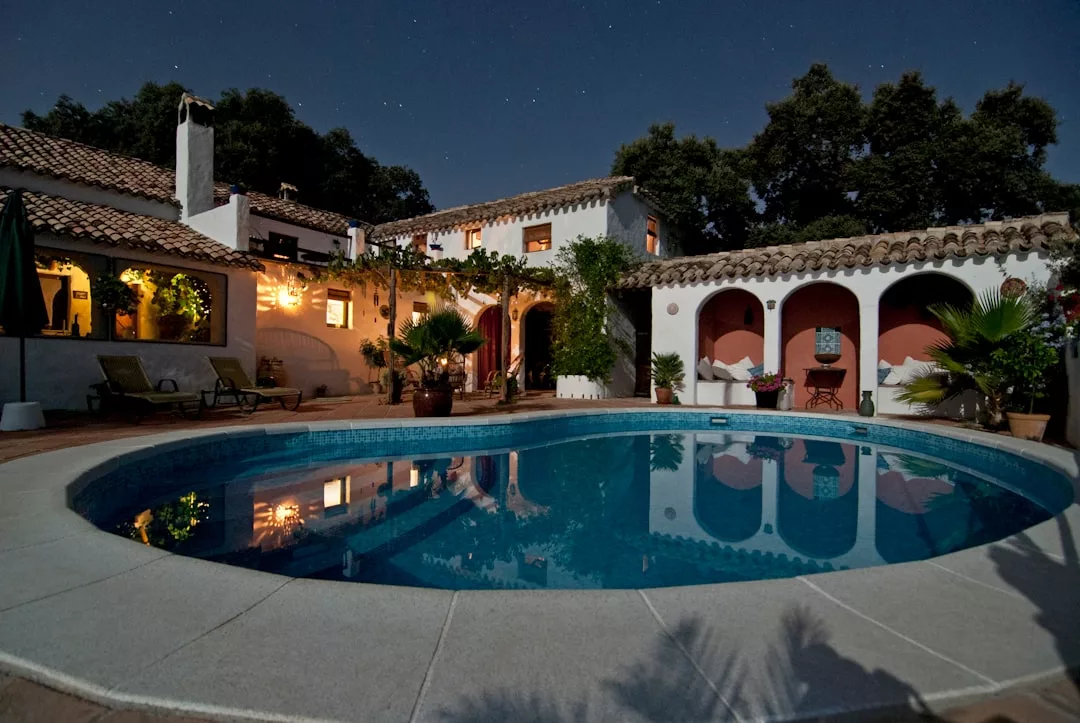 A round swimming pool is surrounded by a luxury Mediterranean-style villa with arched columns and outdoor lighting at night. Trees and stars are visible in the background.