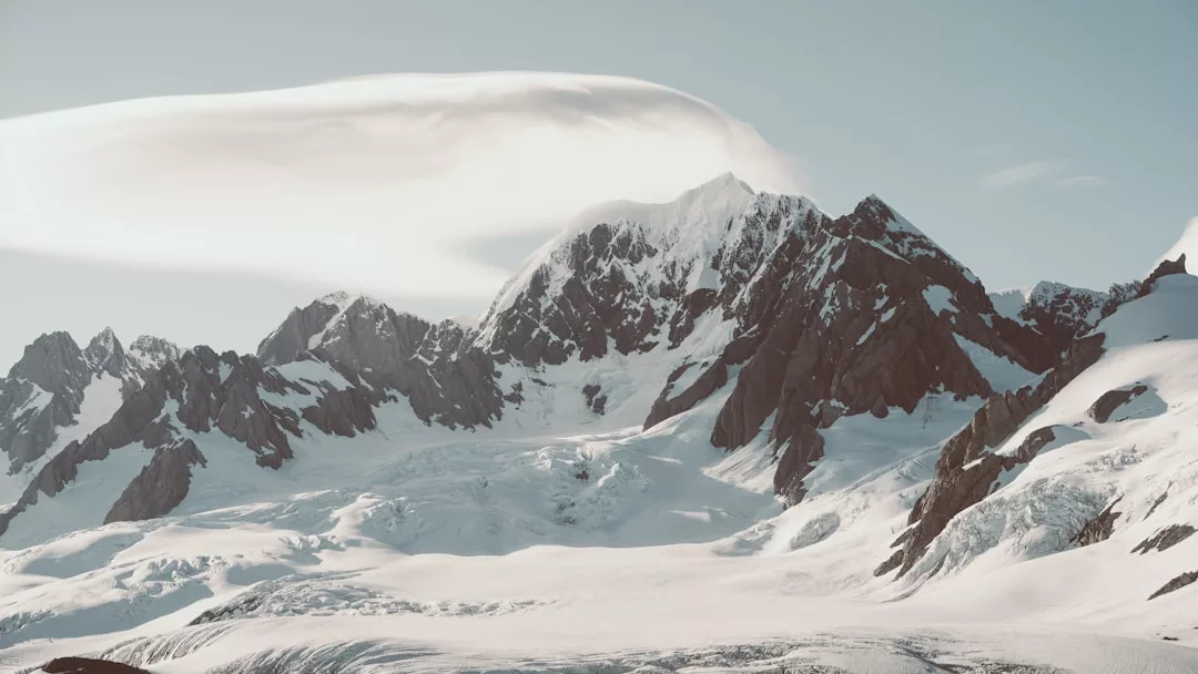 Snow-covered mountain peaks with rugged ridges rise under a clear sky, crowned by a long, curved cloud formation above the highest summit—a scene evoking the pristine luxury of Glamesia.