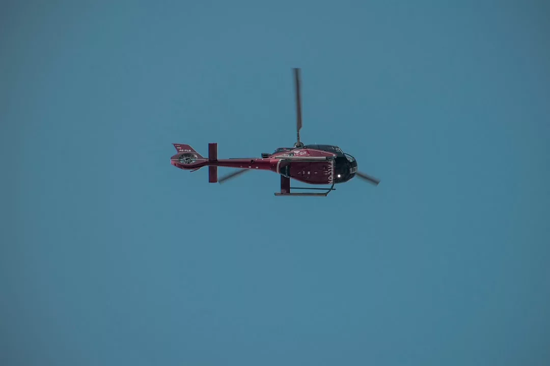 A red helicopter, symbolizing a luxury lifestyle, soars through the clear blue sky, viewed from below.
