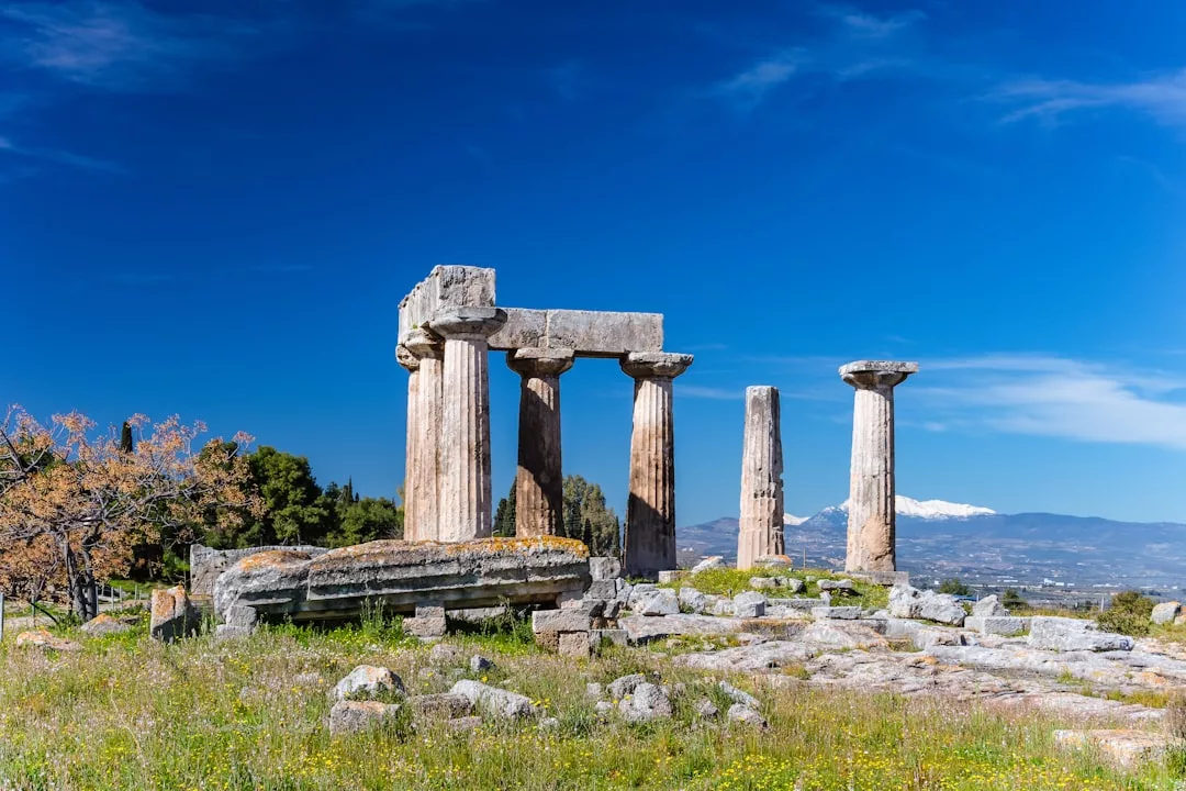 Ruins of ancient stone columns from a Greek temple stand amid grass and scattered stones, with mountains in the background under a clear blue sky—a timeless scene where classical luxury meets natural beauty.
