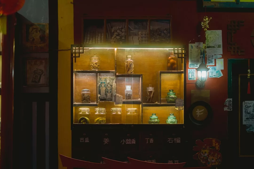 A dimly lit display case with glass jars and bottles containing herbs or ingredients, set against a red and yellow wall adorned with decorations and pictures, evokes a sense of luxury and refined lifestyle.