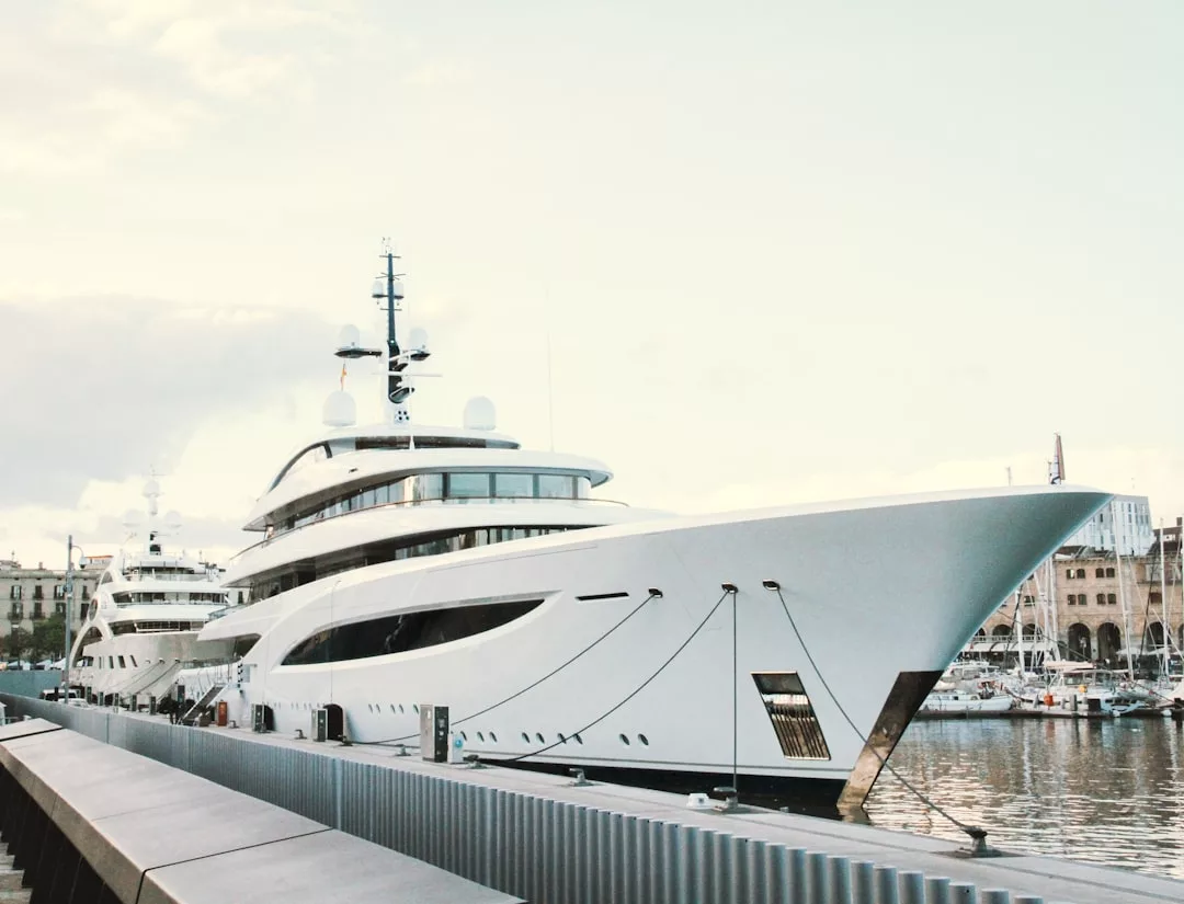 A large white luxury yacht is docked at a marina, with another yacht behind it and buildings in the background, reflecting an upscale lifestyle.