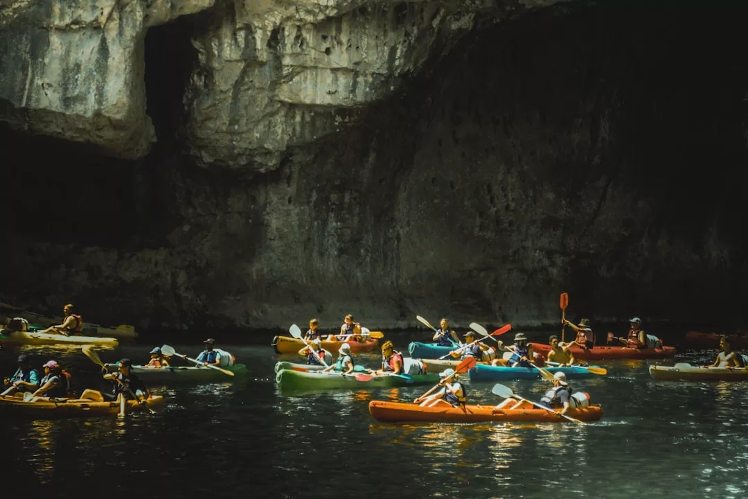 A group of people in colorful kayaks paddle on dark water near a large rocky cave wall, embracing an adventurous lifestyle.
