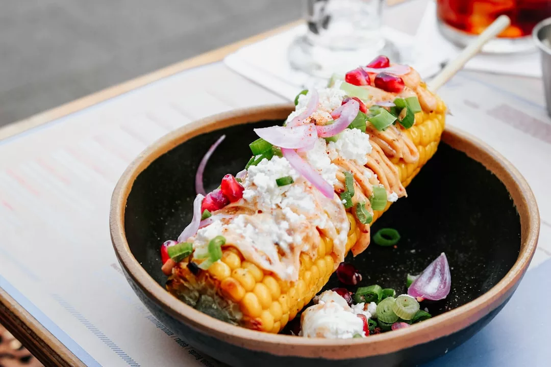 A grilled corn on the cob topped with cheese, sliced red onion, green onions, sauce, and pomegranate seeds is served in a black bowl for a hint of luxury in every bite.