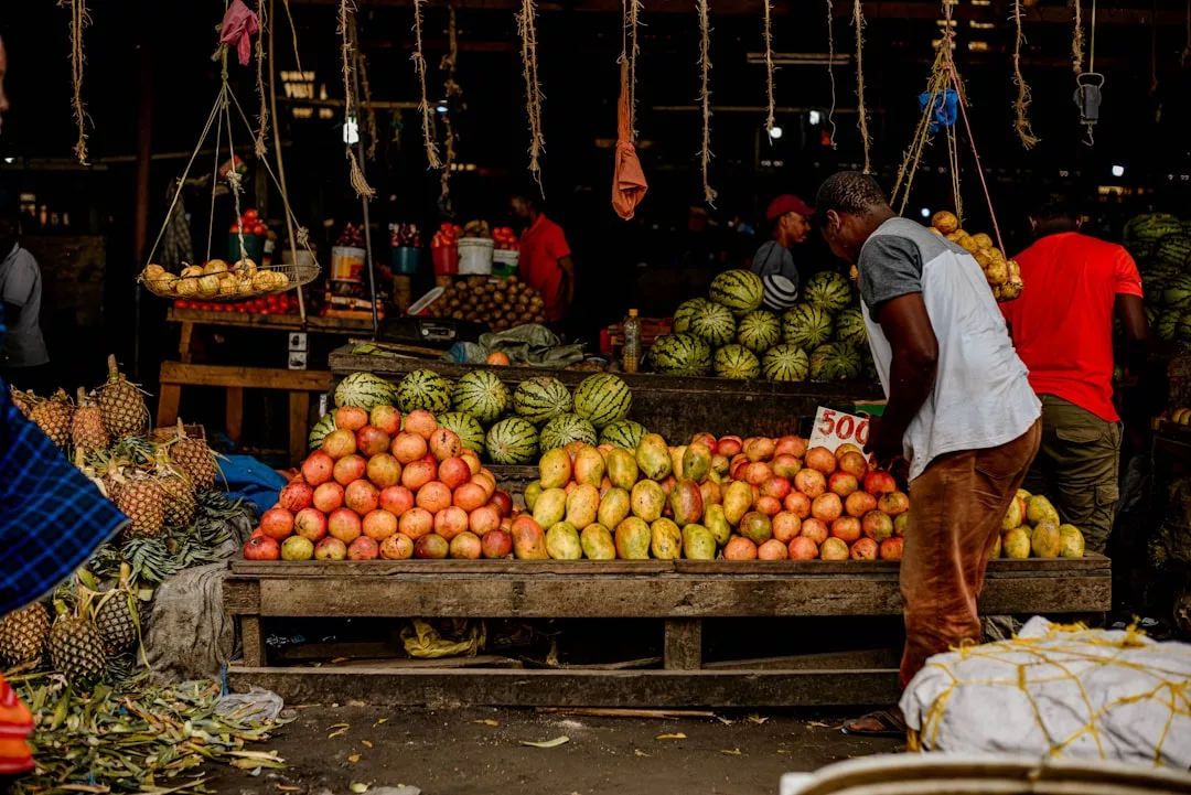 A man arranges piles of mangoes and papayas at a fruit market stall, with watermelons and pineapples visible in the background, capturing the vibrant lifestyle of fresh produce markets.