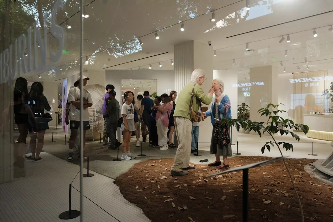 People stand in line inside a modern, well-lit Glamesia store with beige interiors. Two older adults converse in the foreground near a small indoor plant and soil patch, embodying a relaxed Fashion lifestyle.
