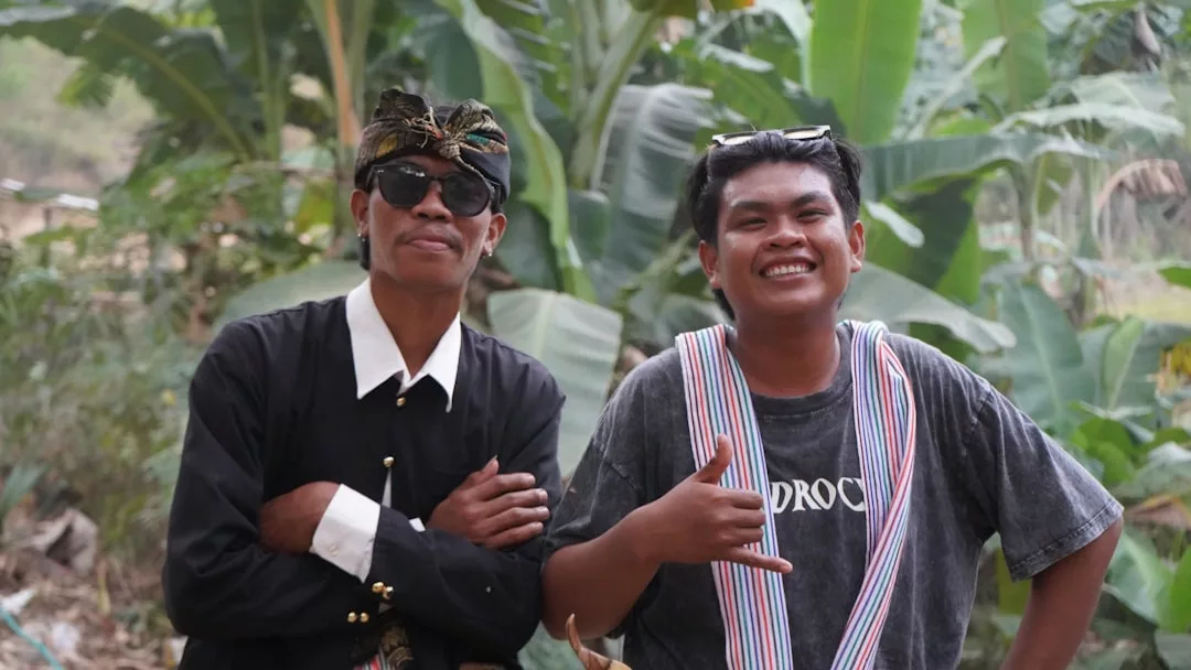 Two men stand outdoors in front of lush green plants; one showcases luxury fashion with traditional attire and sunglasses, while the other wears casual clothing and smiles, making a hand gesture.