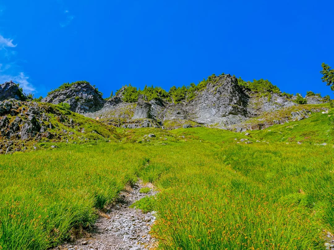 A grassy trail leads up a green hillside toward rocky cliffs under a clear blue sky, creating a picturesque scene perfect for a lifestyle escape.