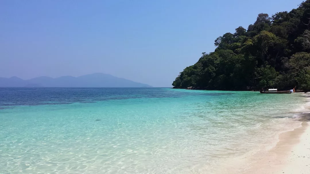Clear turquoise water meets a sandy beach with a forested hill on the right, under a blue sky and distant mountains—a scene of pure luxury and tranquility.