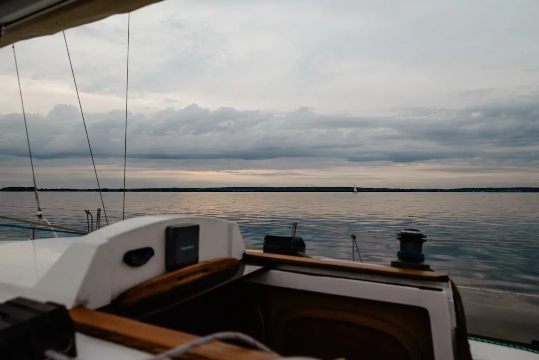 Experience the luxury lifestyle aboard a sailboat, gliding over calm water beneath a cloudy sky, with a distant shoreline and a small sailboat accenting the horizon.