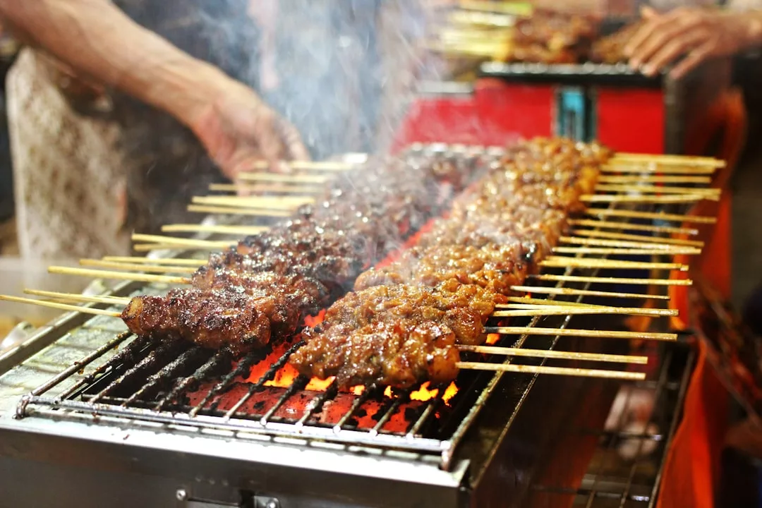 Several skewers of meat are being grilled over open flames on a street food barbecue, with smoke rising and hands tending the skewers, capturing the vibrant Glamesia lifestyle.