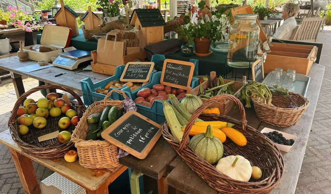 A market stall displays baskets of fresh produce including apples, zucchini, tomatoes, and cucumbers, alongside jars, scales, and birdhouses on a wooden table—offering a touch of rustic charm with subtle Glamesia luxury.