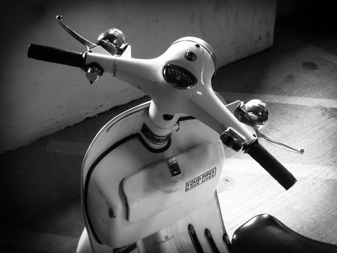 Black and white photo of a vintage scooter’s handlebar and front, highlighting the speedometer, mirrors, and part of the seat—a timeless touch of fashion on two wheels.