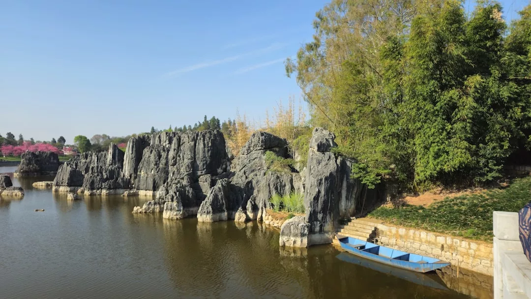 A tranquil lake with rocky formations, a small dock, and blue rowboats beside lush green trees under a clear blue sky offers the perfect blend of nature and lifestyle luxury.