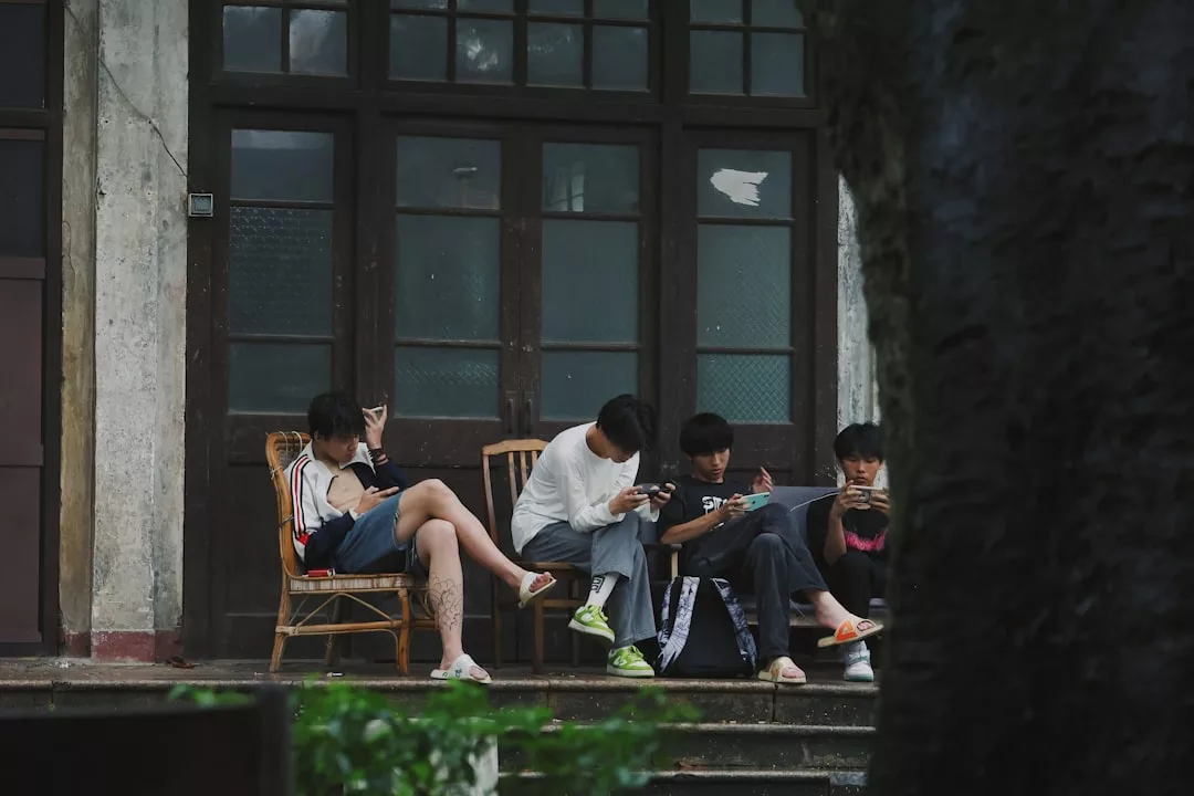 Four people sit on chairs outside a building, each focused on their phones or tablets, enjoying a relaxed lifestyle amid greenery and a tree in the foreground.
