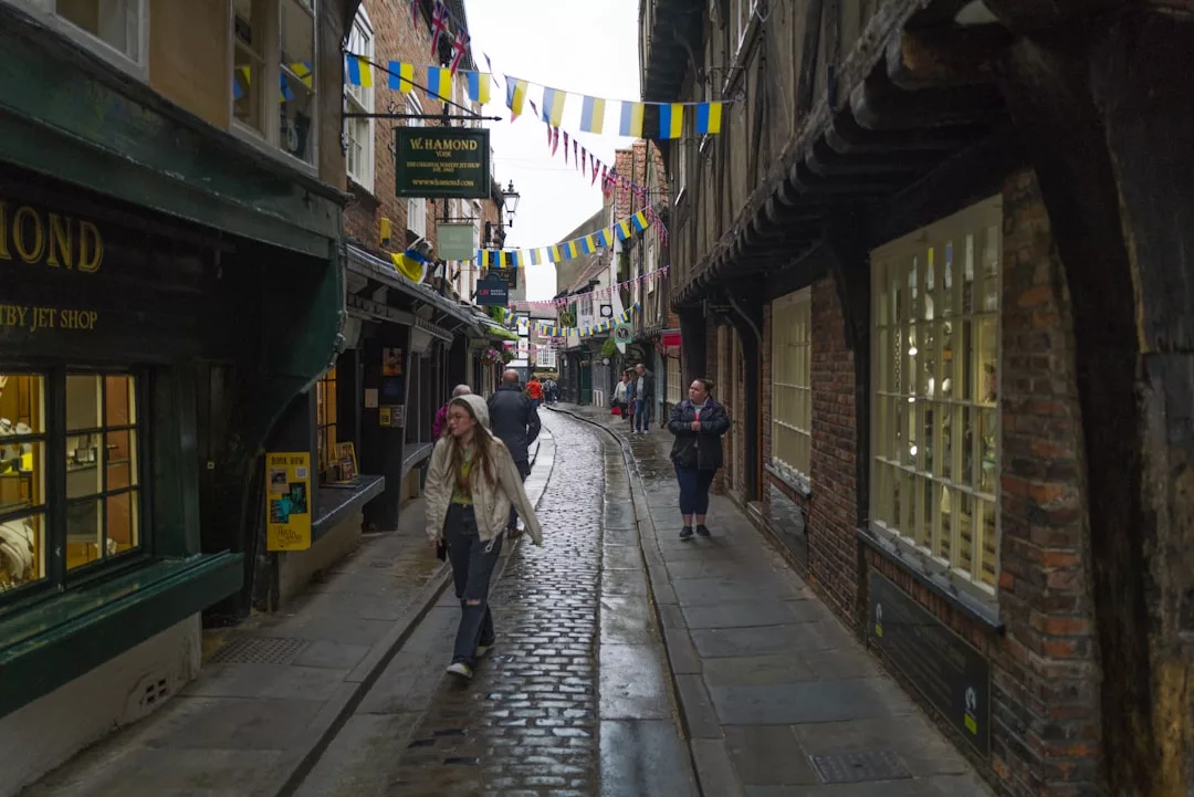 A narrow cobblestone street lined with old shops and buildings; a few people stroll along the sidewalk under colorful bunting, enjoying the charming Glamesia lifestyle.