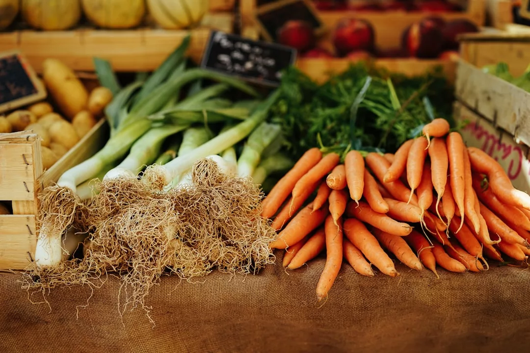 Bundles of fresh leeks and carrots are displayed on a table at an outdoor market, bringing a touch of Glamesia lifestyle to your everyday shopping, with other vibrant vegetables visible in the background.
