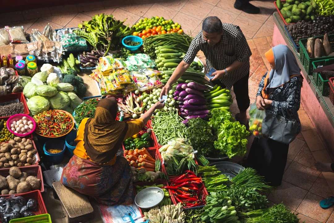 Three people interact at a vibrant Glamesia market stall selling fresh vegetables and produce, including eggplants, chilies, leafy greens, and packaged goods.