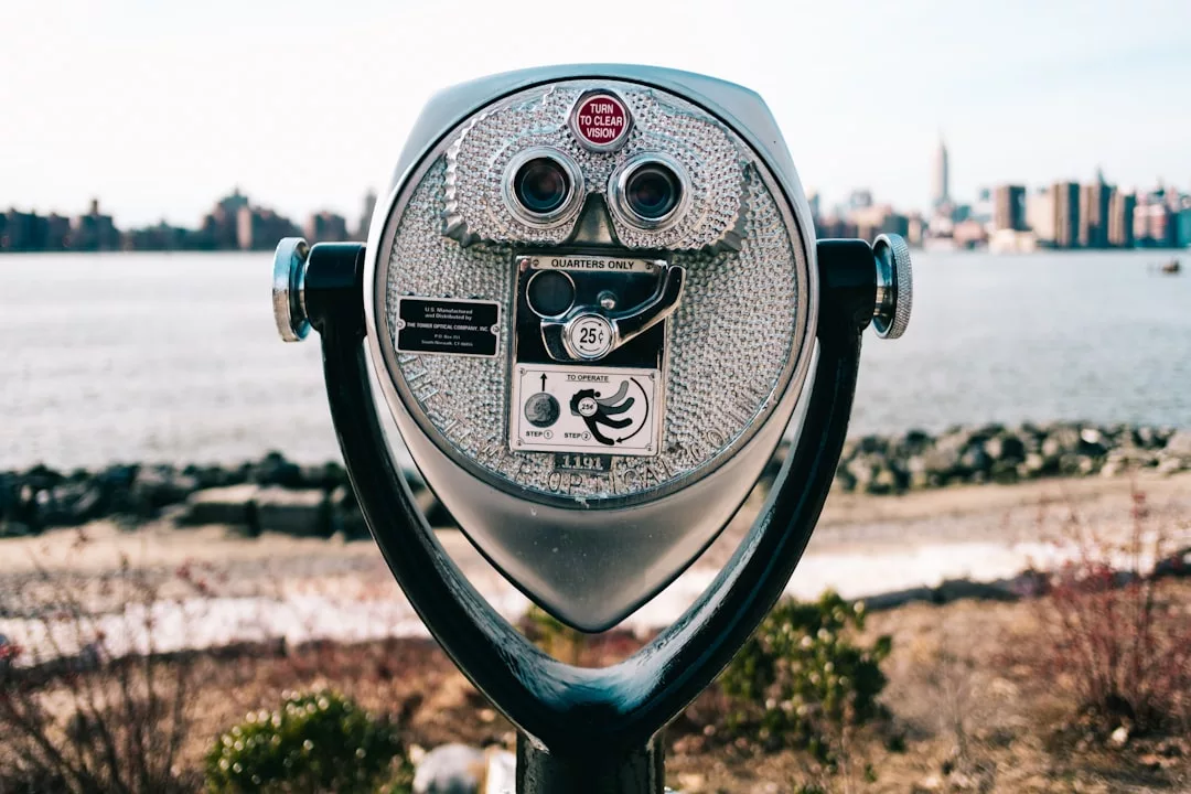 A coin-operated binocular viewer is positioned outdoors, overlooking a body of water and a city skyline in the background, offering a glimpse of urban luxury.