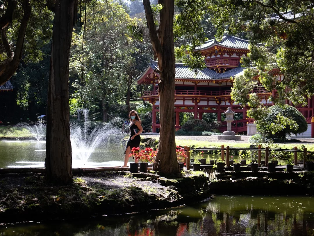 A person wearing a mask stands by a pond and fountain in a garden with traditional Japanese architecture and lush trees, exuding an air of luxury and fashion.